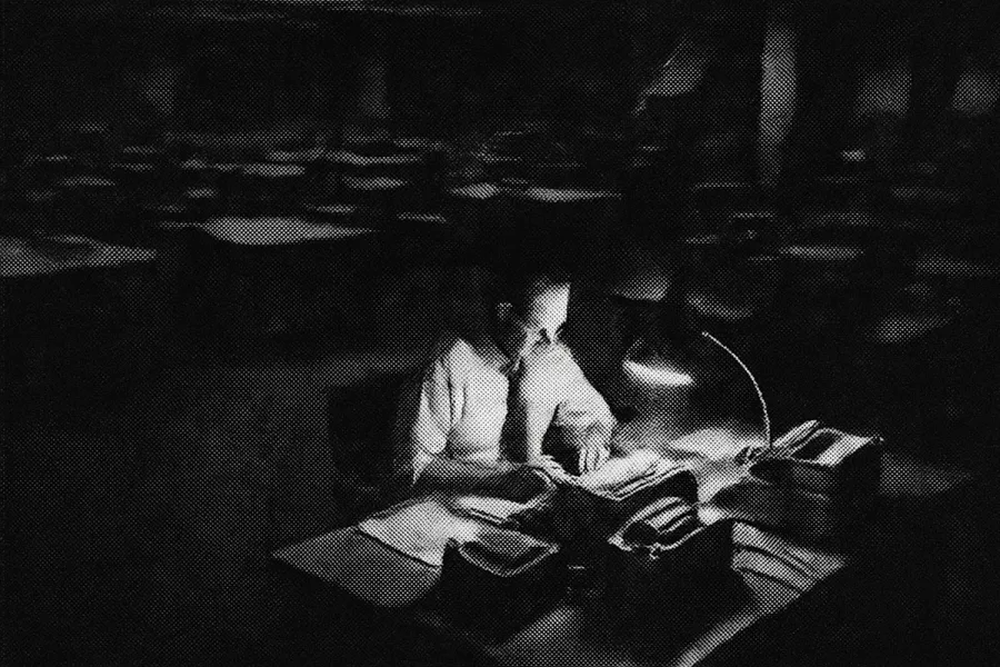 One man at a lamp-lit desk in a vast empty newsroom, halftone photo